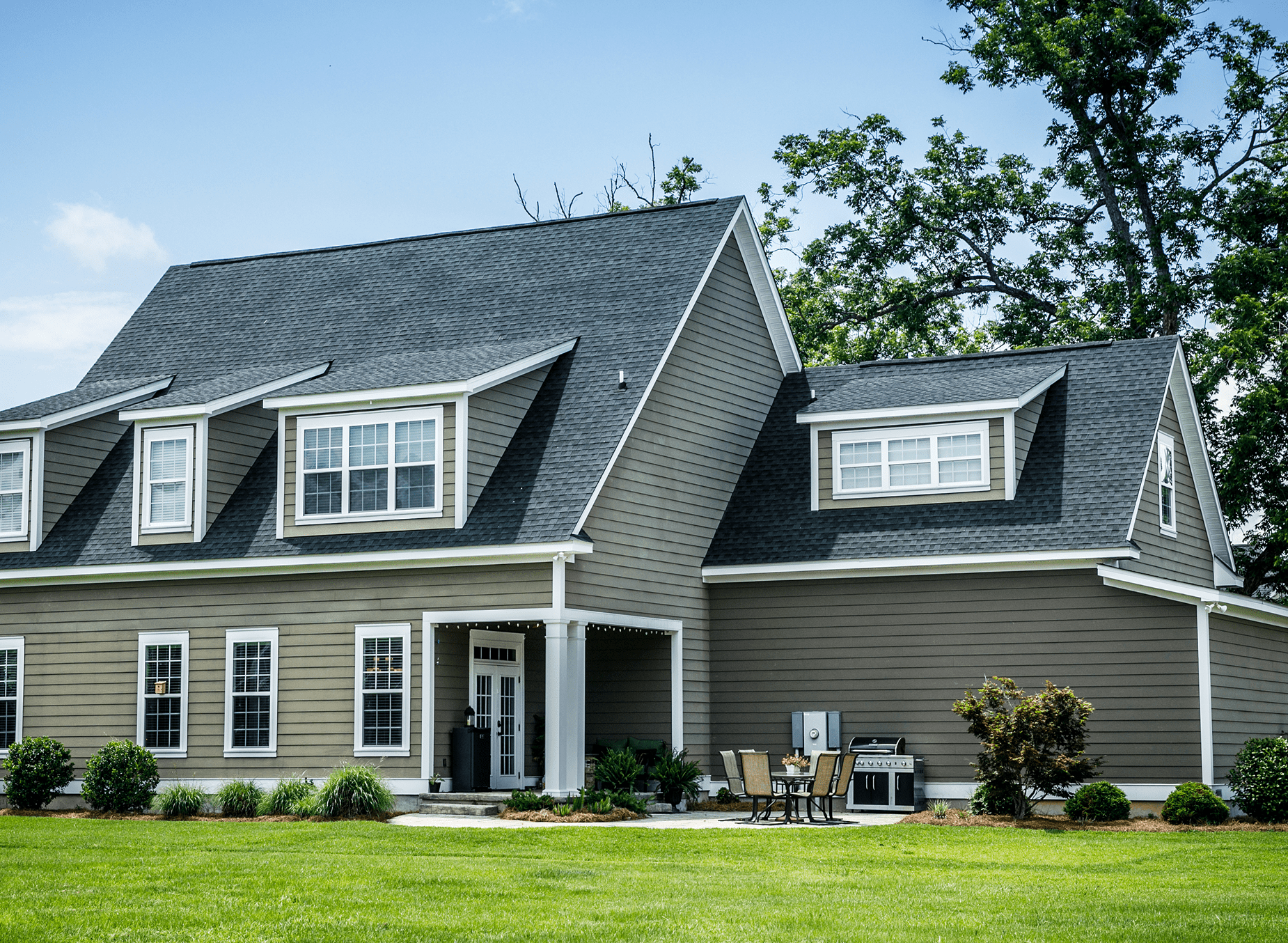 Rectangle 1142 A large, gray two-story house with dormer windows and a well-maintained lawn. A patio set and grill are on the side. Bright, sunny day with a few trees around.
