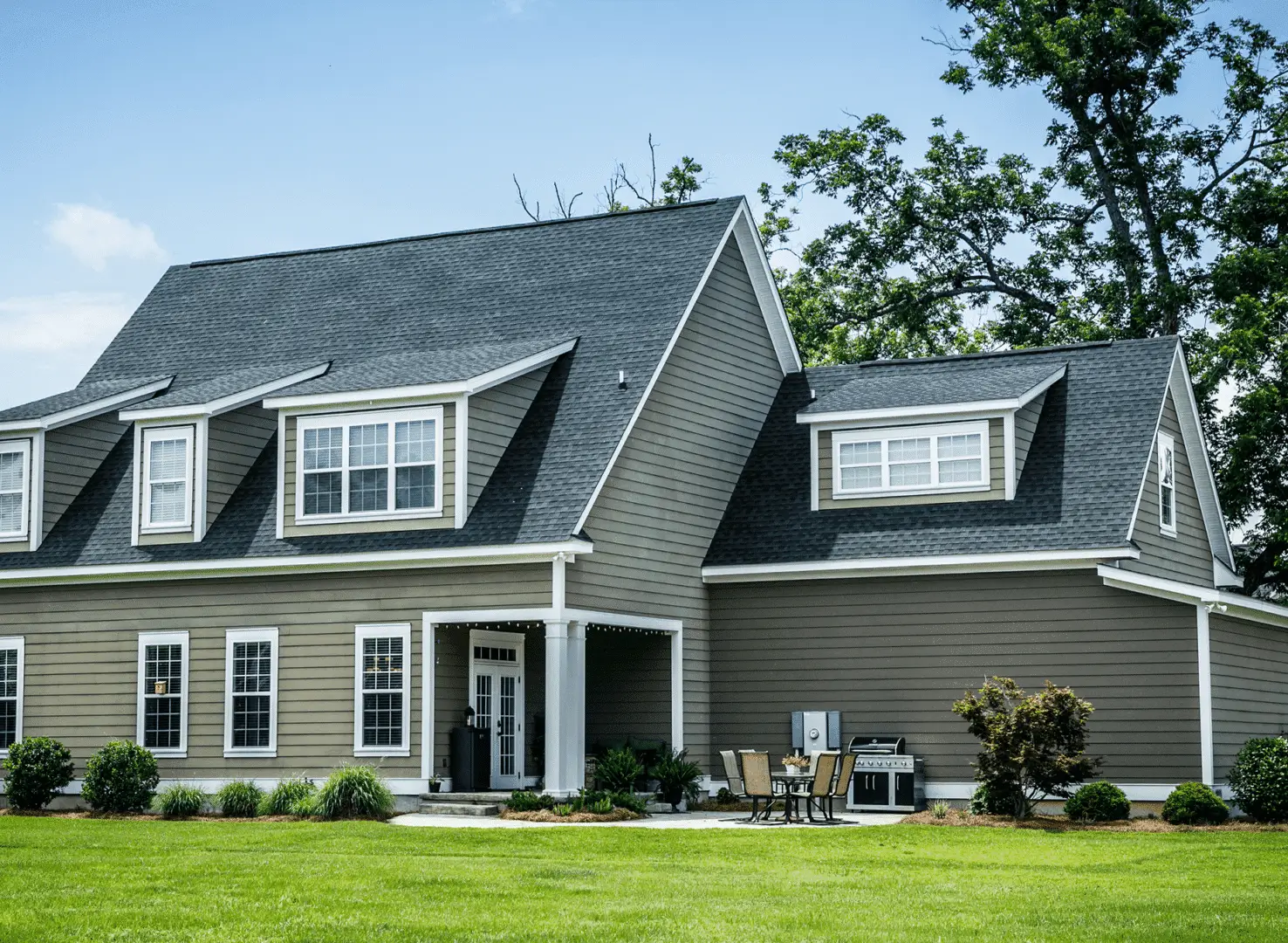A large, gray two-story house with dormer windows and a well-maintained lawn. A patio set and grill are on the side. Bright, sunny day with a few trees around. A large, gray two-story house with dormer windows and a well-maintained lawn. A patio set and grill are on the side. Bright, sunny day with a few trees around.